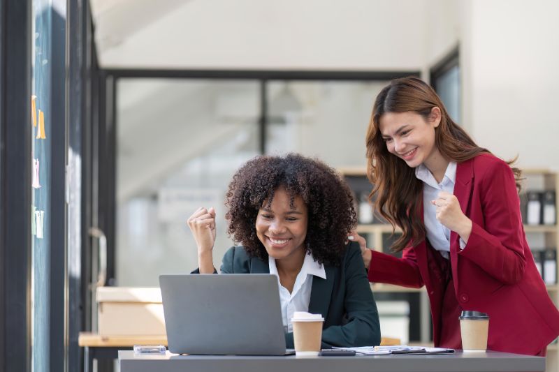 Two businesswomen celebrating AP automation success while working on a laptop in office setting.